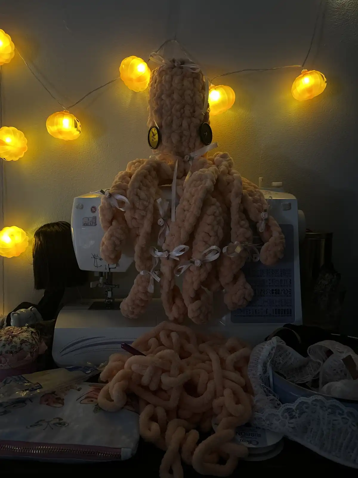 A large, pink, chunky yarn crocheted octopus wearing headphones sits atop a white sewing machine in a dimly lit room. Warm…
