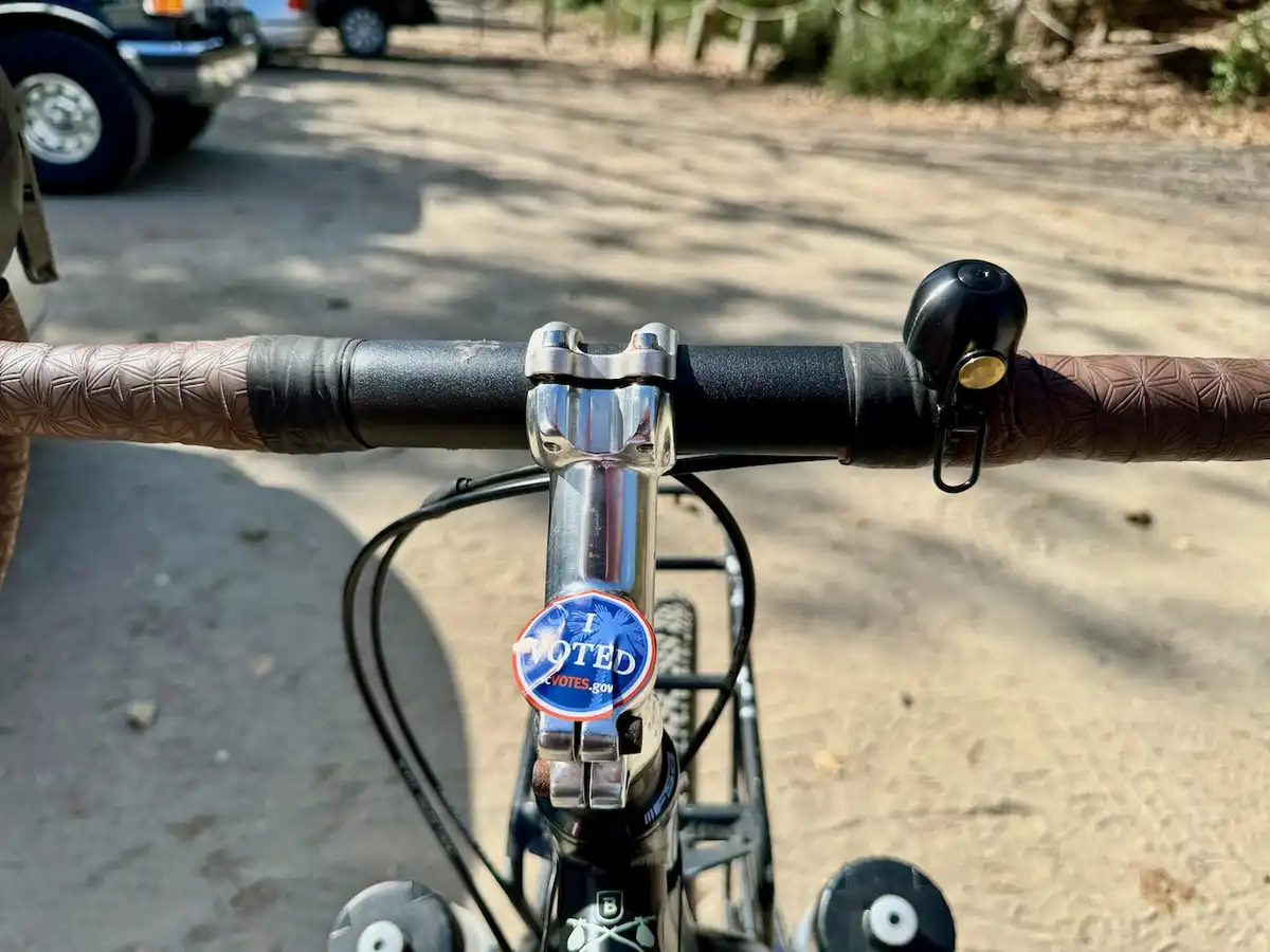 A close-up photo from a cyclist's perspective shows a blue circular 'I VOTED' sticker attached to the chrome stem of a…