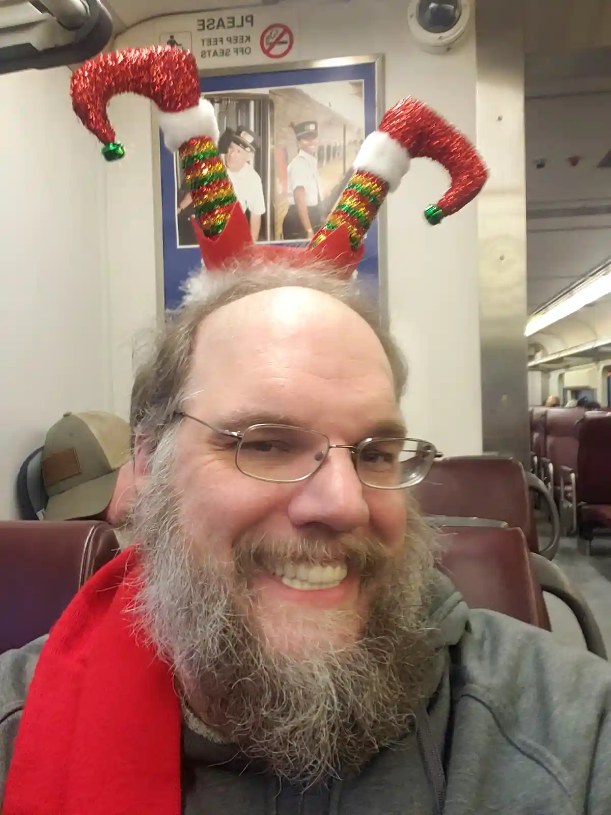A man with a gray beard and glasses smiles while wearing a red, glittery elf hat with jingle bells on a train. He wears a…