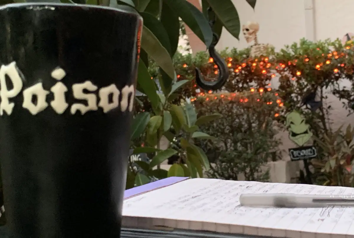 A black pint glass with the word 'Poison' in white gothic lettering is in the foreground, sitting on a notebook with a pen…
