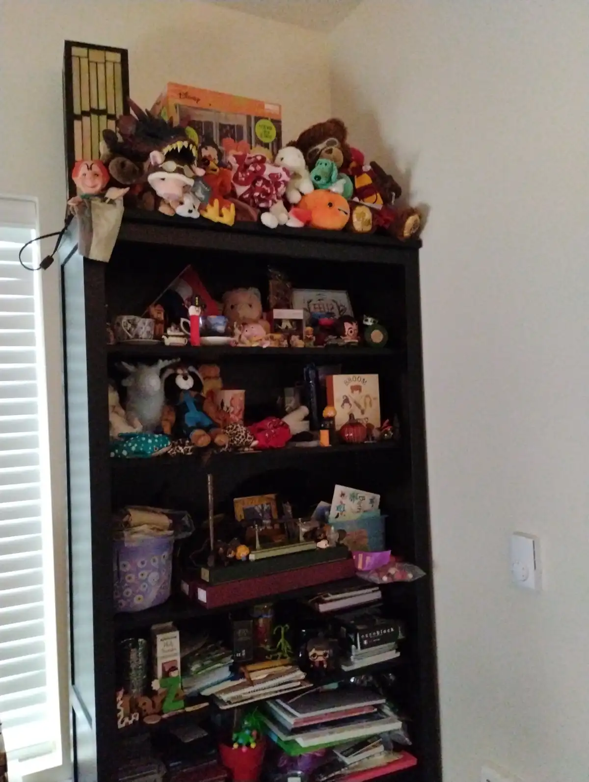 A dark wooden bookshelf is completely overflowing with clutter, including stuffed animals, books, toys, and miscellaneous…