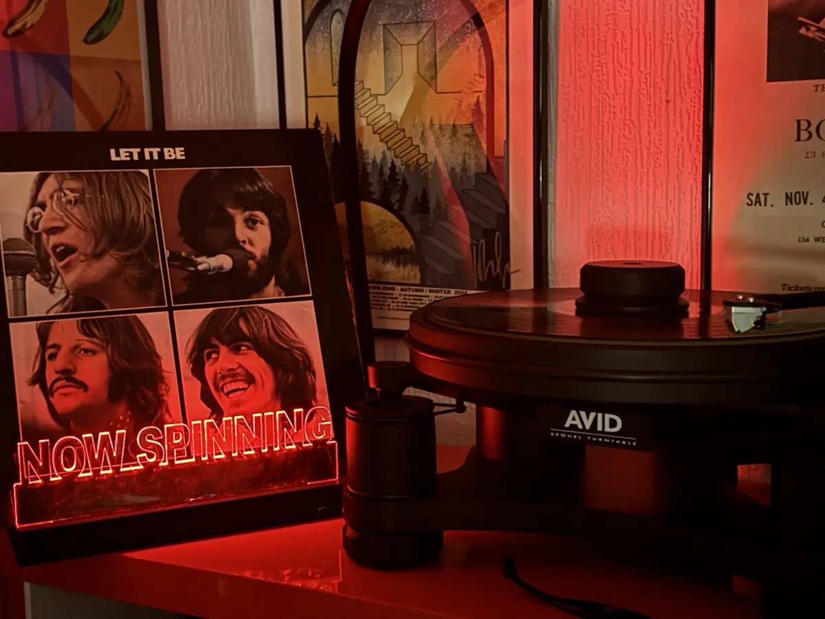A red-lit room features a vinyl record player with a record on it, next to a framed 'Let It Be' album cover showing the four…