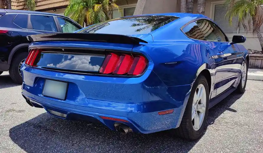 A shiny blue Ford Mustang is parked outdoors, its rear view showing red taillights, a spoiler, and a blurred license plate…