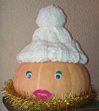 A pumpkin is decorated with googly eyes, pink lips, and a gold tinsel beard, topped with a white knit hat and pom-pom. The…