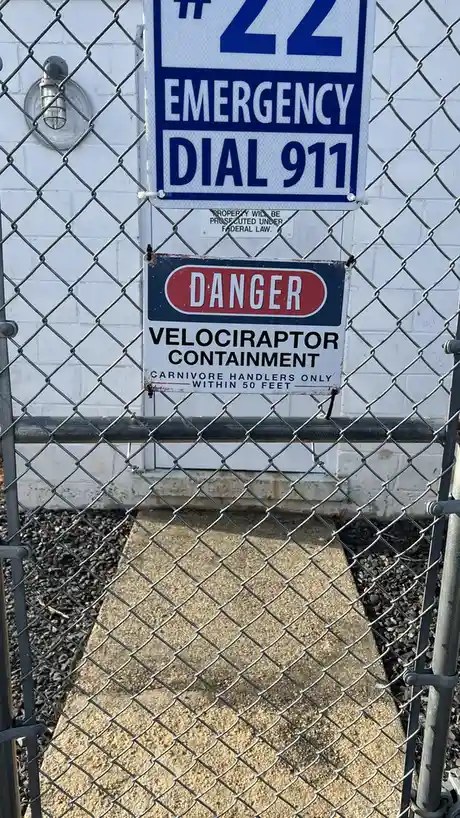 A chain-link fence encloses a white brick wall with two signs. The top sign reads "#22 EMERGENCY DIAL 911". Below it, a…