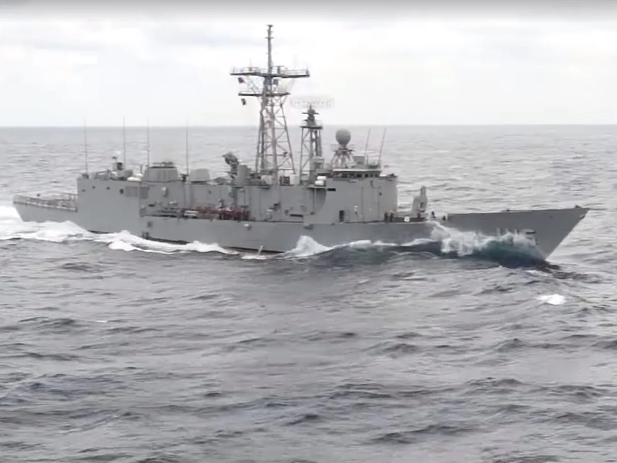 A gray naval warship is shown sailing through choppy ocean waters, its bow cutting through waves. The vessel is equipped…