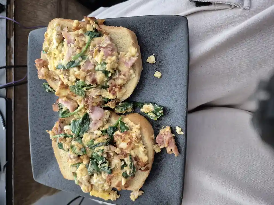 A close-up photo shows a messy, open-faced breakfast sandwich on a dark gray plate, with scrambled eggs, bacon, and spinach…