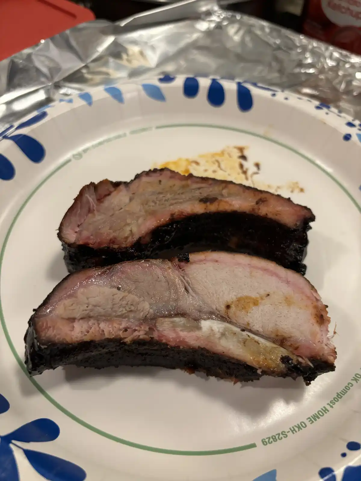 Two slices of smoked pork butt are on a white paper plate with blue and green patterns, showing a dark charred exterior and…