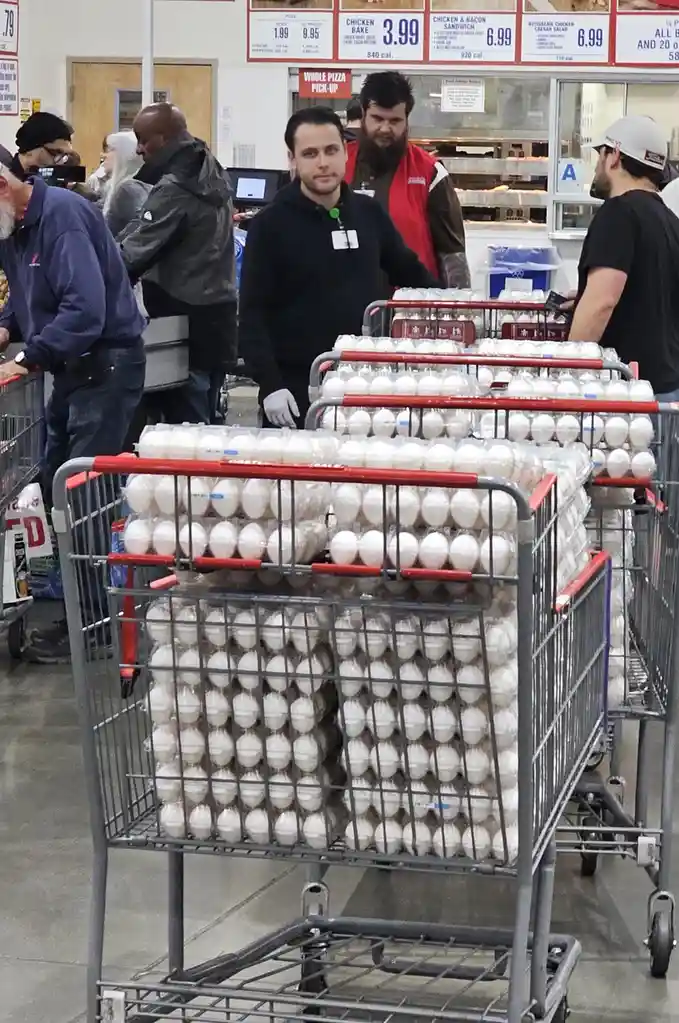 A man in a black hoodie and white gloves stands in a grocery store aisle, looking directly at the camera with a neutral…