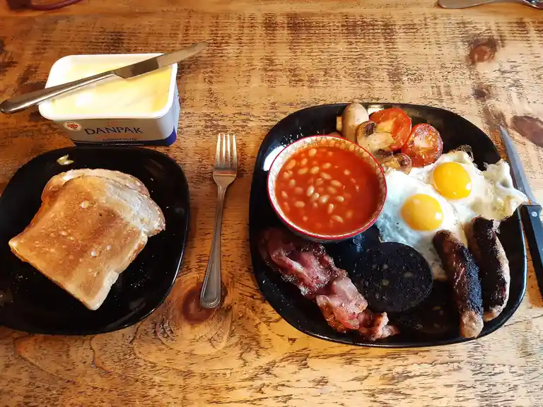 A hearty full english breakfast is laid out on a wooden table, featuring fried eggs, bacon, sausages, black pudding, baked…