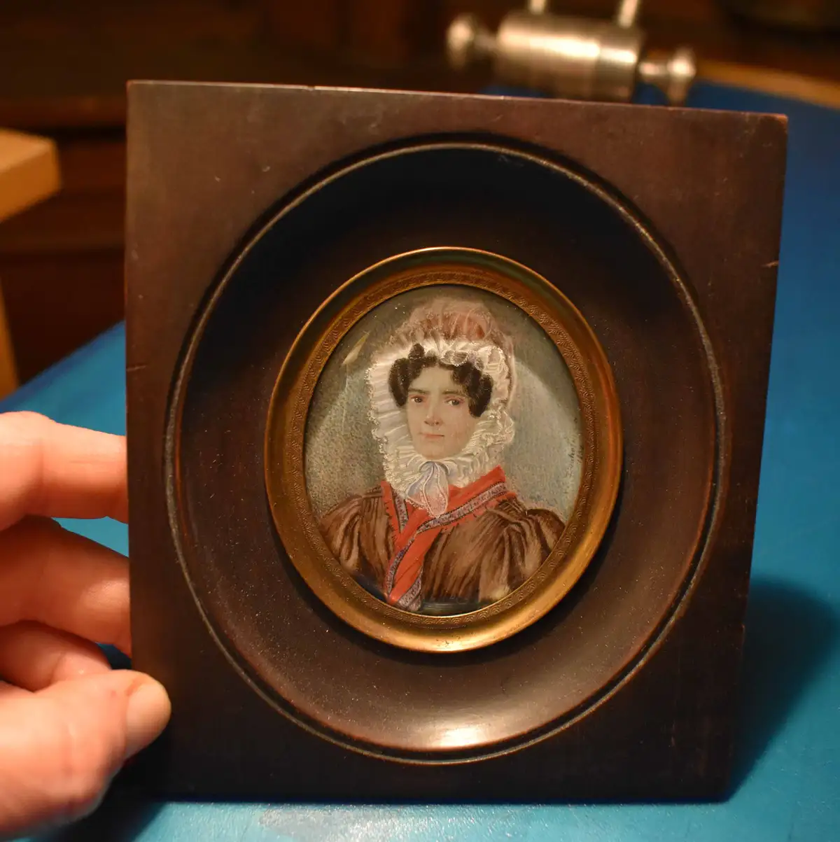 A hand holds a dark wooden-framed miniature portrait of a woman in 19th-century attire, featuring a white lace bonnet and…