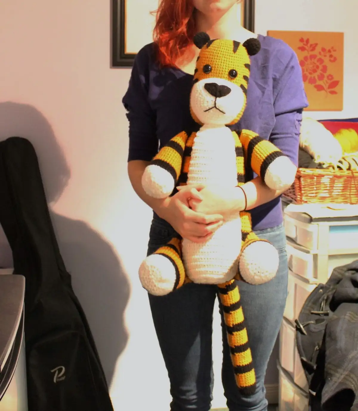 A person with red hair, wearing a purple shirt and jeans, holds a large, handmade crocheted tiger with orange and black…