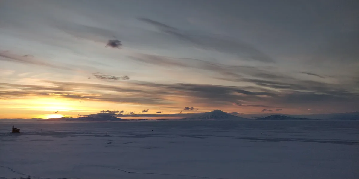 A wide, serene landscape photo of a snowy, frozen plain under a dramatic sunset sky with distant mountains. The sun is low…