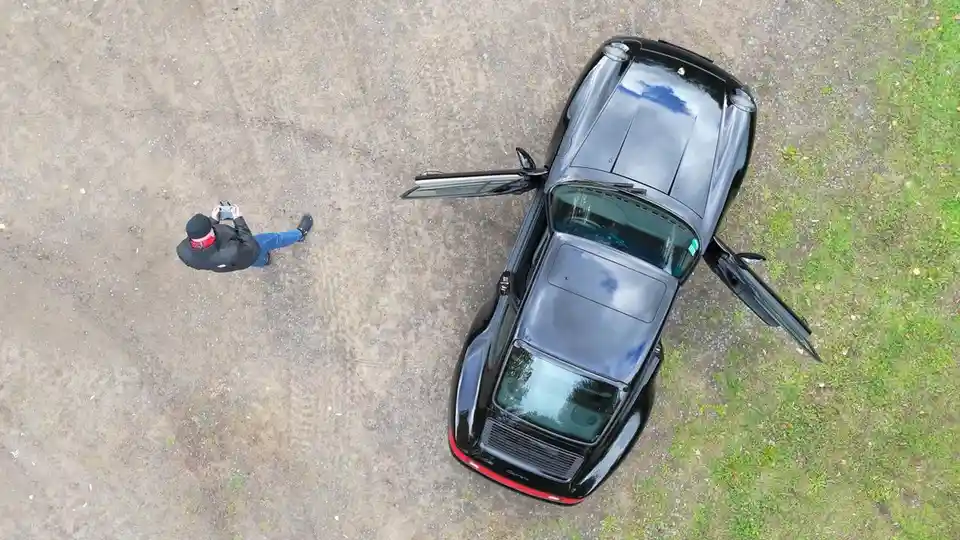 An overhead shot shows a person in a black jacket and blue jeans standing on dirt ground, holding a phone and looking at a…