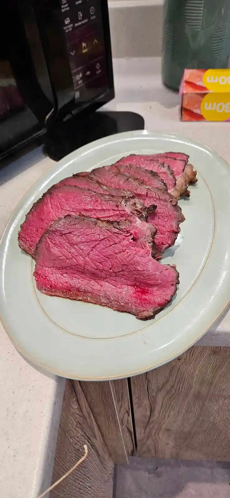 A plate of sliced medium-rare steak with a vibrant pink center sits on a kitchen counter, with a microwave and a green water…