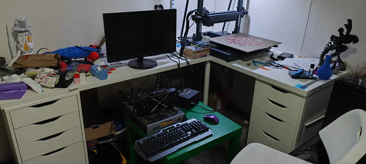 A cluttered desk setup with a computer monitor, keyboard, mouse, and a partially assembled PC motherboard on a green table…