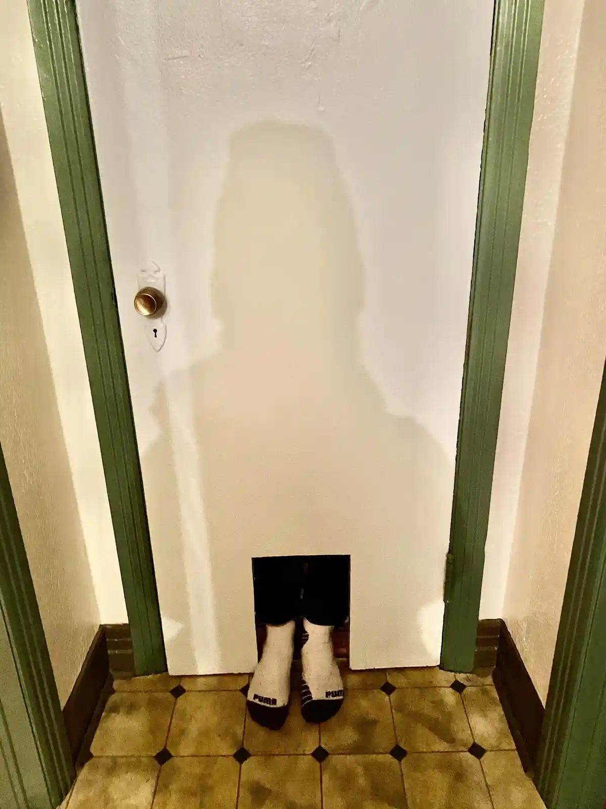 A person's shadow is cast on a white door, but only their feet in white Puma socks are visible through a rectangular hole…