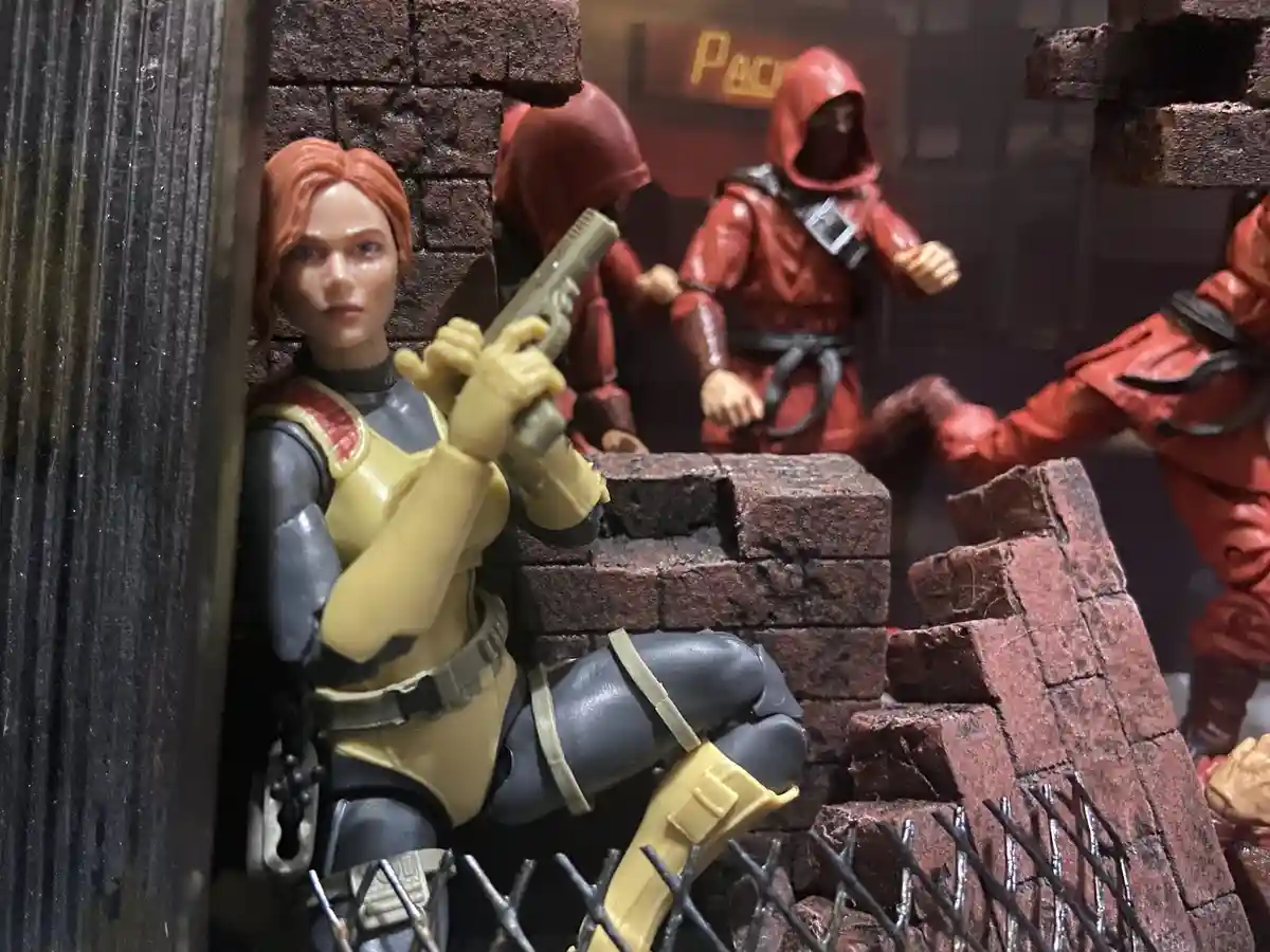 A toy action figure of a woman with red hair, wearing a yellow and grey outfit and holding a pistol, crouches behind a brick…