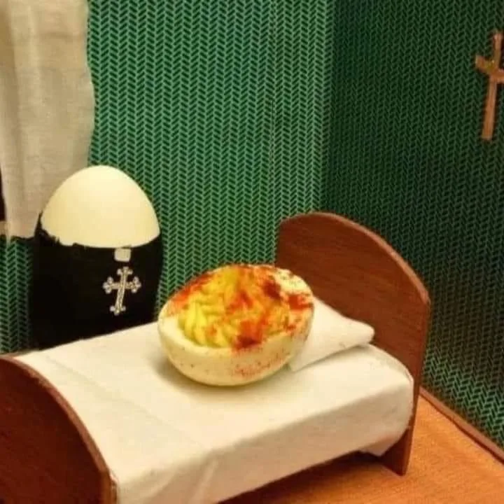 A large, hollowed-out egg filled with macaroni and cheese sits on a miniature bed with a white sheet and pillow. Behind it…