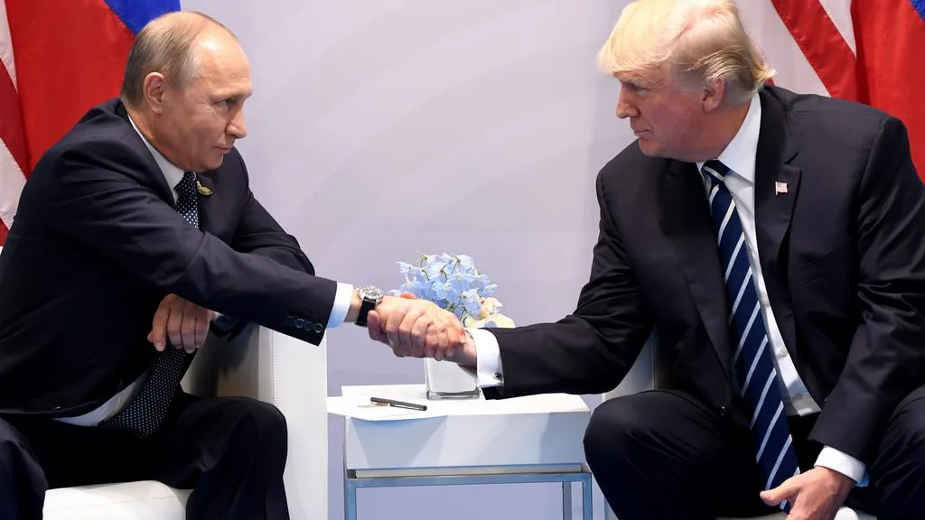 Two men in suits, identified as Vladimir Putin and Donald Trump, are seated and shaking hands across a small table with a…