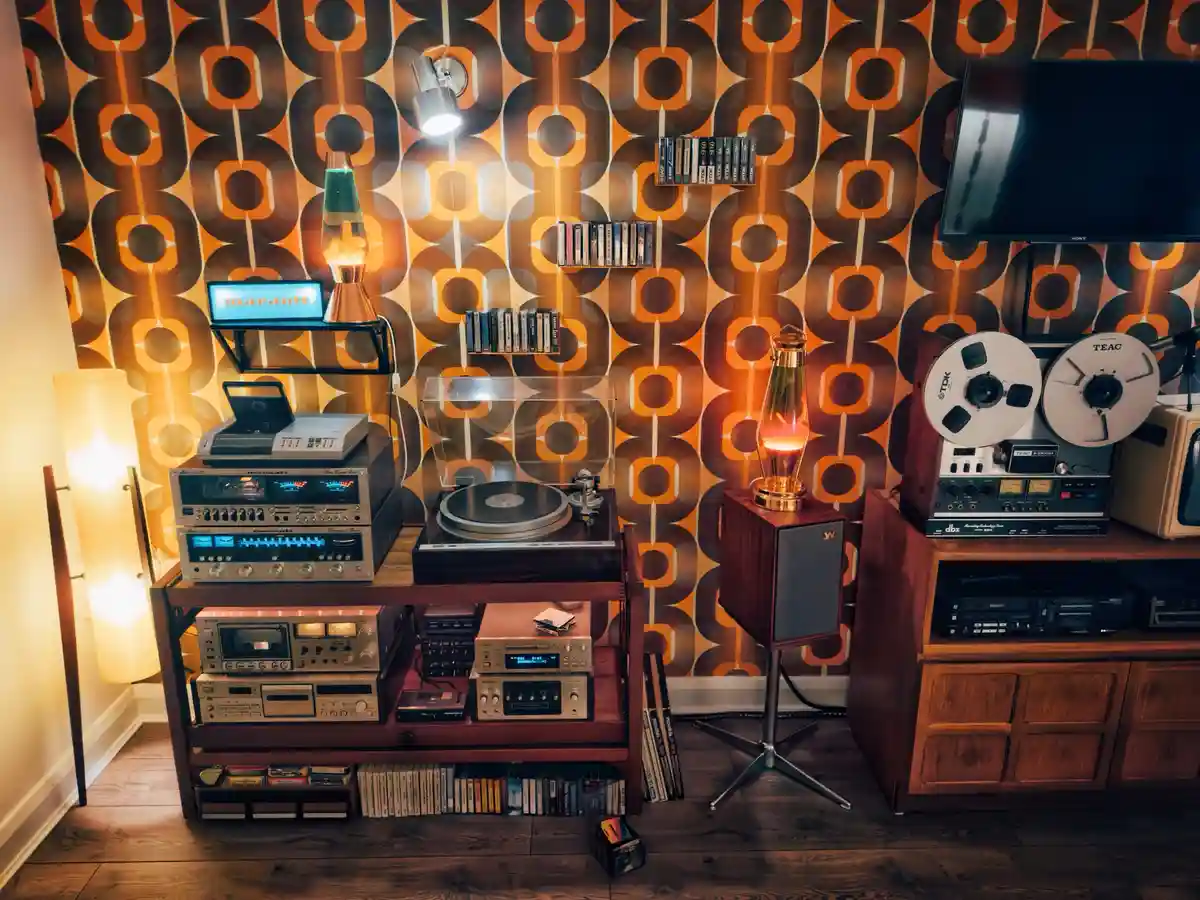 A warmly lit room with retro orange and brown patterned wallpaper features a large wooden stereo system filled with vintage…
