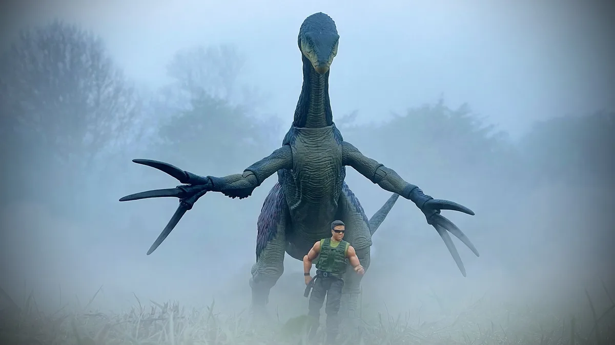 A small action figure of a man in tactical gear stands in front of a large, menacing dinosaur action figure in a foggy…