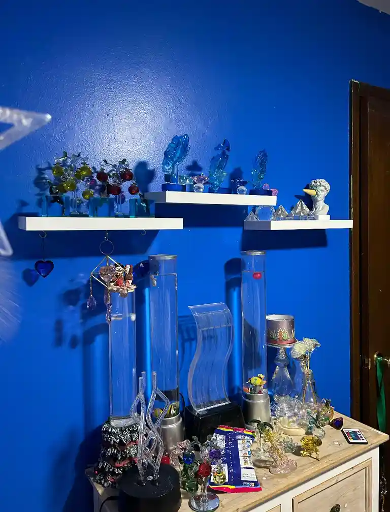 A room with bright blue walls features white shelves holding numerous glass figurines, sculptures, and decorative items…