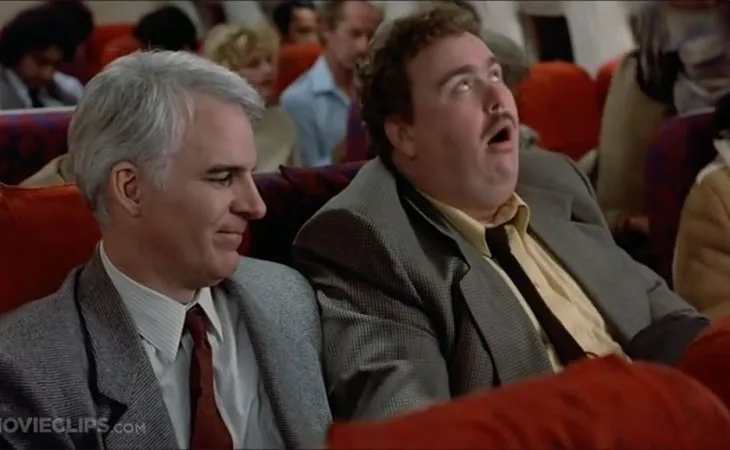 Two men sit side by side in a theater; the man on the right has a shocked, open-mouthed expression while the man on the left…