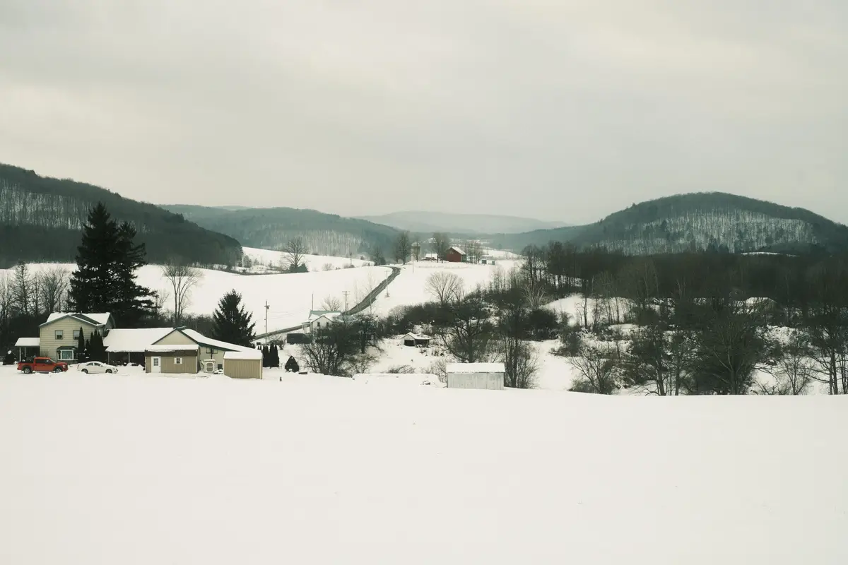 A wide, snowy rural landscape with houses, bare trees, and rolling hills under an overcast sky. A red pickup truck and a…