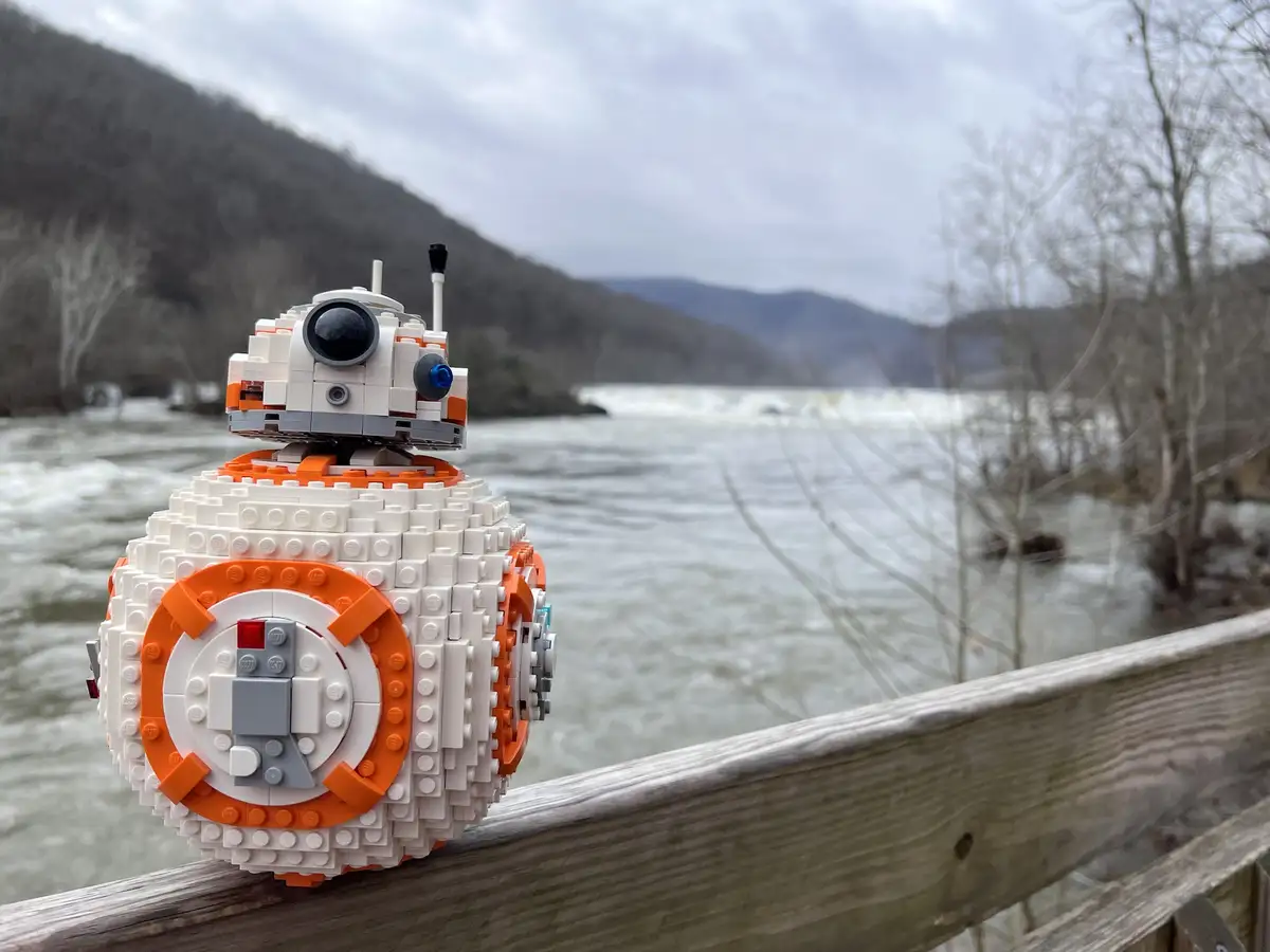 A LEGO model of the Star Wars droid BB-8 sits on a wooden railing overlooking a turbulent river with mountains in the…