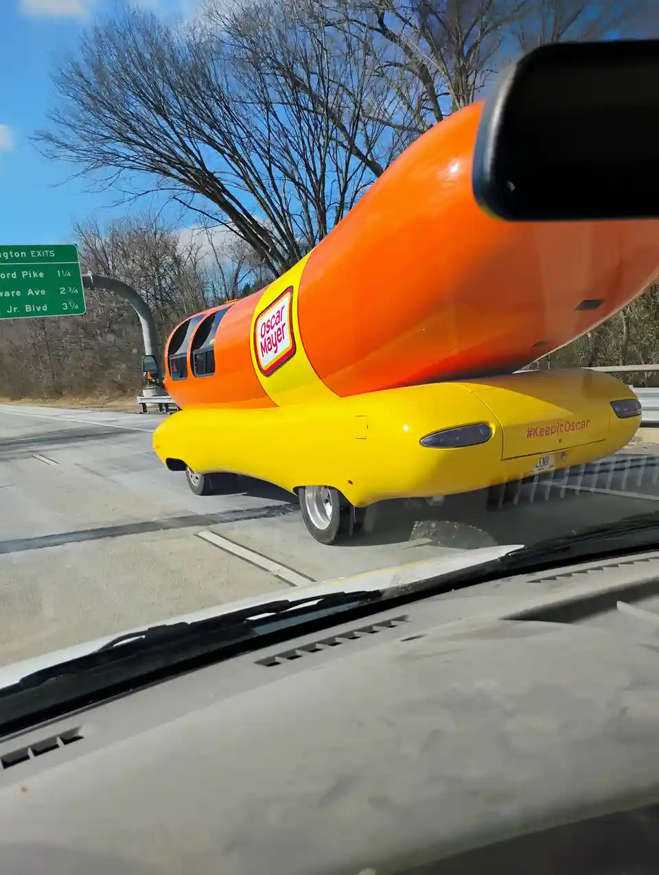A photo taken from inside a car shows the iconic Oscar Mayer Wienermobile, shaped like a giant hot dog with orange and…