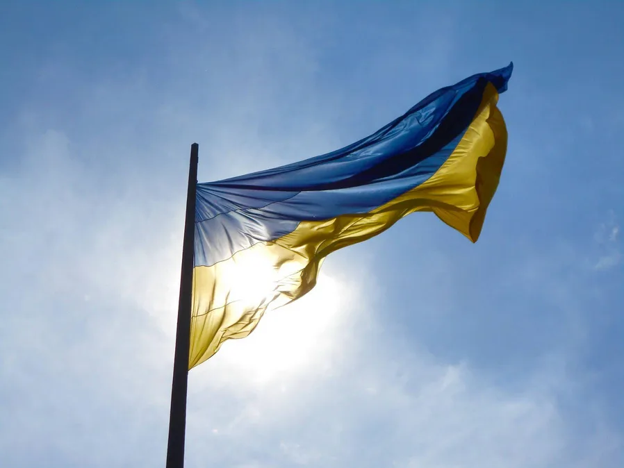 A Ukrainian flag with blue and yellow horizontal stripes waves in the wind against a bright blue sky with the sun shining…