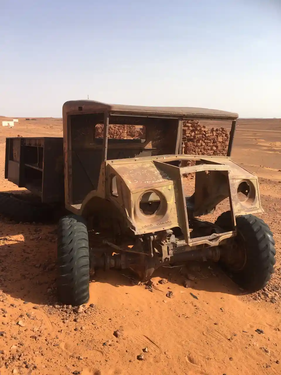 a rusted, stripped-down, old ford canada truck sits abandoned in a barren desert landscape with orange sand and distant…