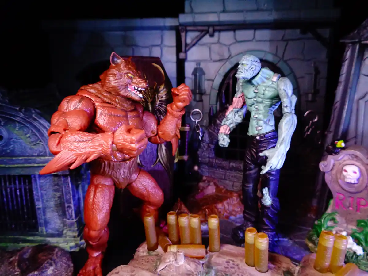 Two action figures, one red werewolf and one green zombie-like creature, stand in a spooky graveyard diorama with tombstones…
