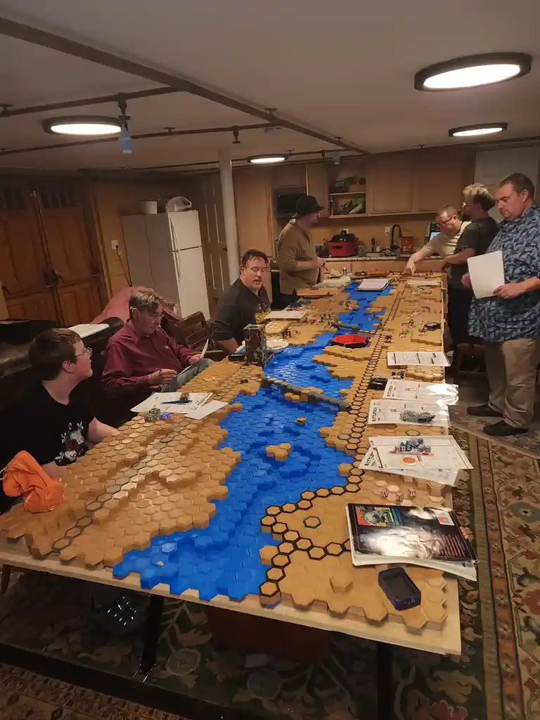 A group of men are gathered around a very long table covered in a massive hexagonal grid map made of tan and blue tiles…