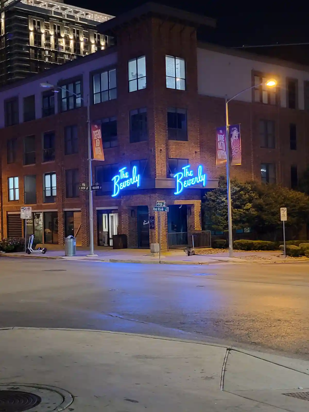 A nighttime street view of a brick building with two identical blue neon signs reading 'The Beverly' mounted on its facade…