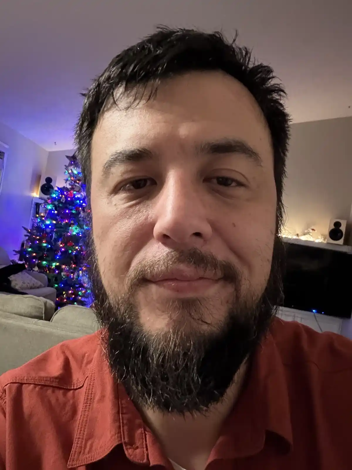 A close-up selfie of a man with a beard and dark hair, wearing a red shirt, looking directly at the camera with a slightly…