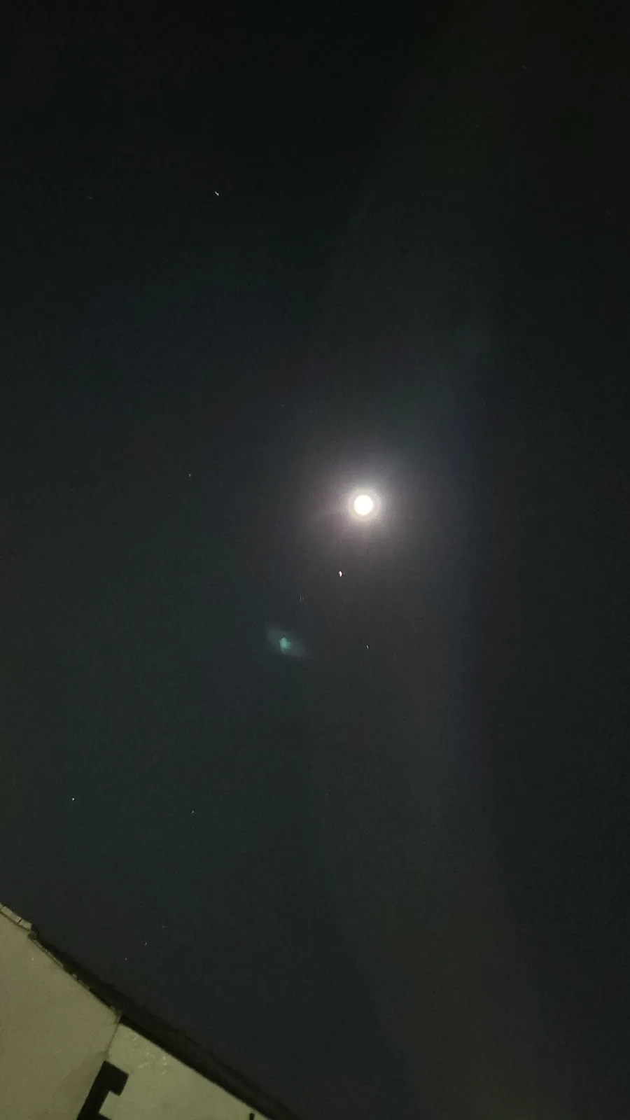 A nighttime photo showing a bright moon in a dark sky with some lens flare and faint stars, with the corner of a building…