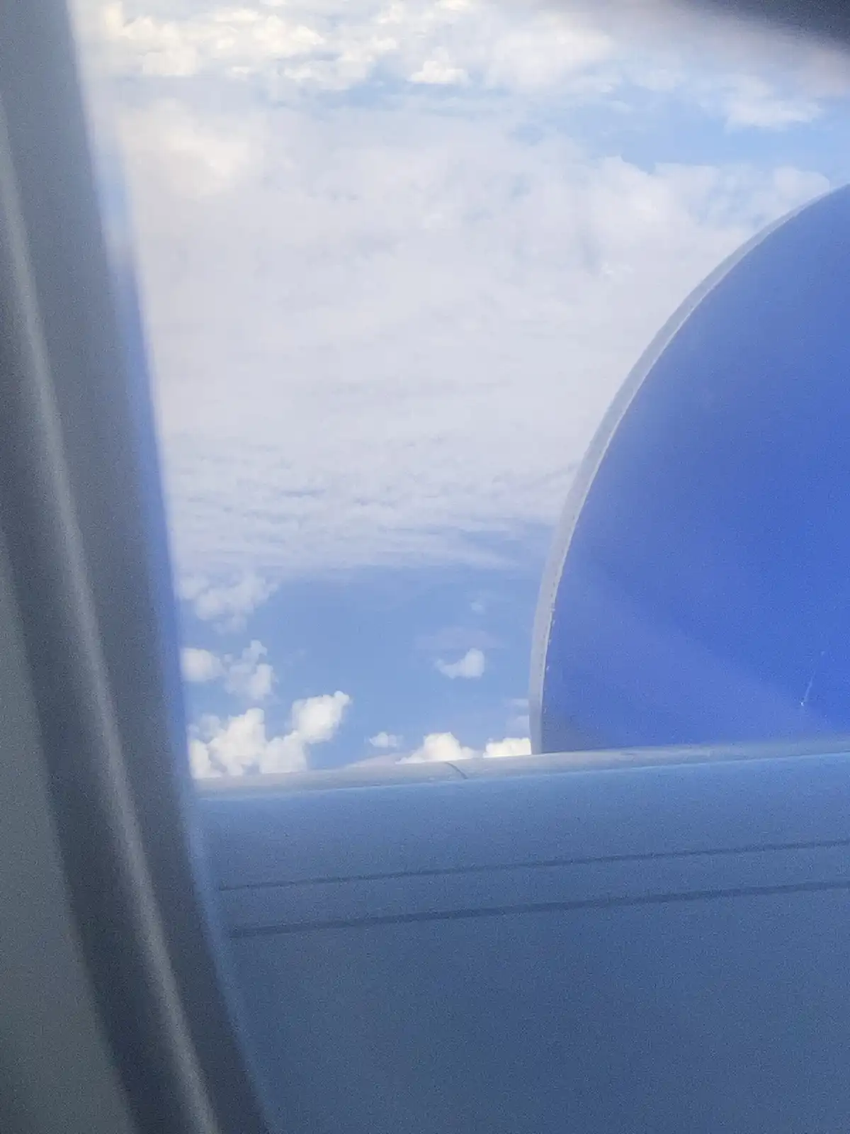 A photo taken from inside an airplane, looking out the window at a cloudy sky. The view is framed by the airplane's window…