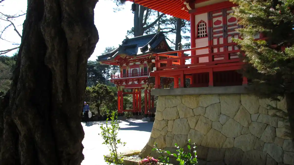A photograph of a traditional Japanese shrine with red wooden structures, a stone wall, and trees, with a person walking in…