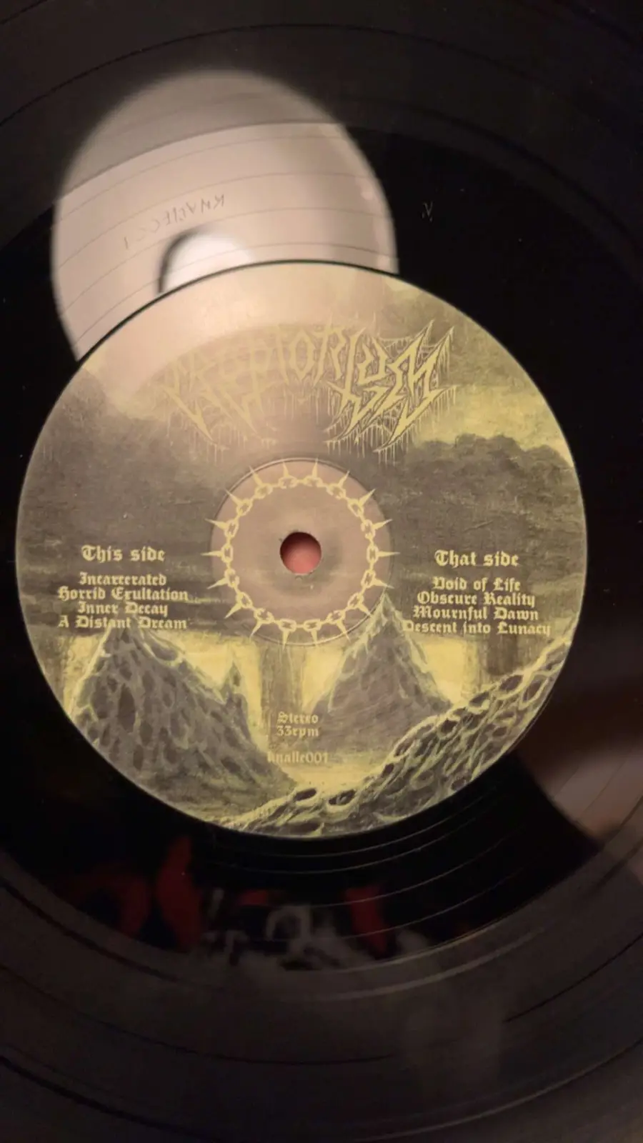 A close-up photo of a black vinyl record label with a dark, textured artwork featuring a spiked chain around the center…