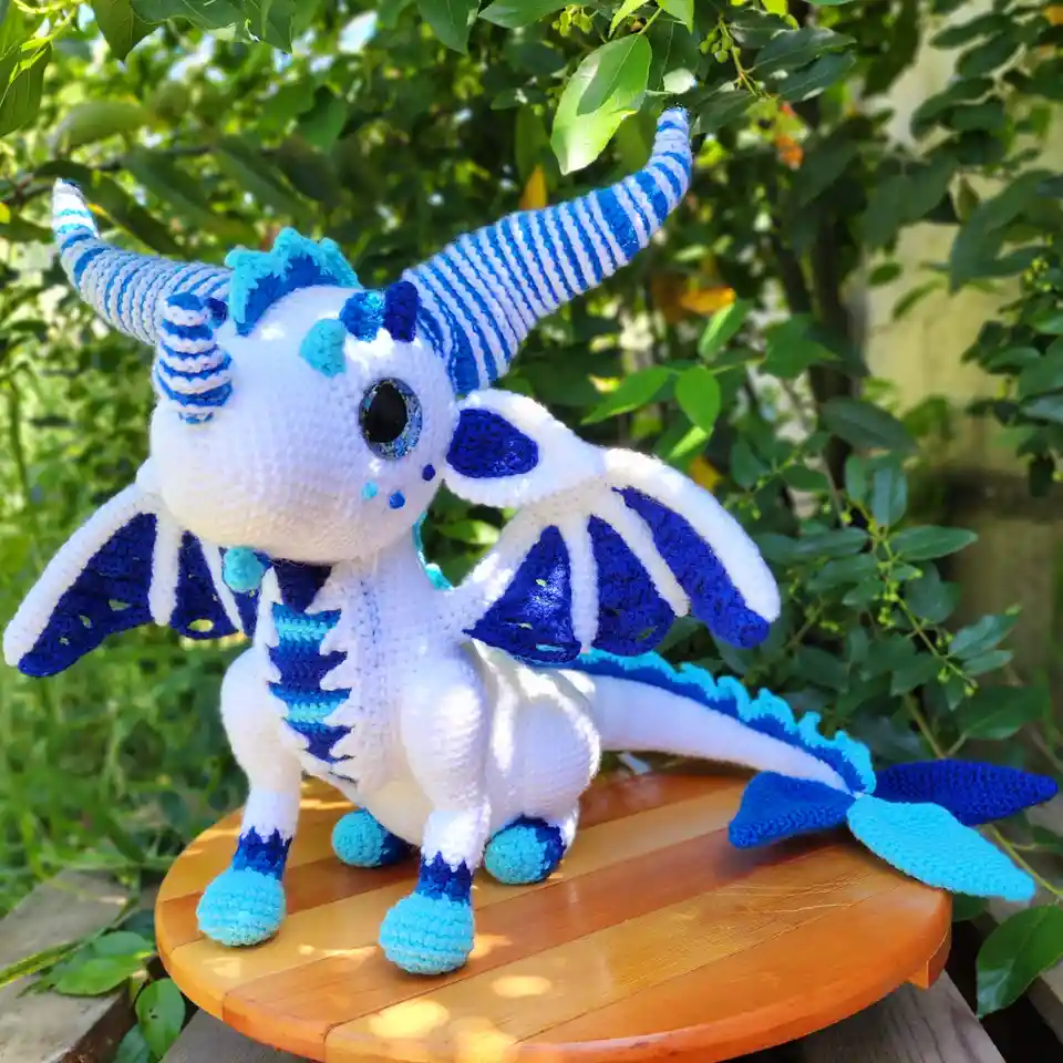 A small, crocheted dragon toy with white and blue yarn, featuring large eyes, striped horns, and wings, sits on a wooden…