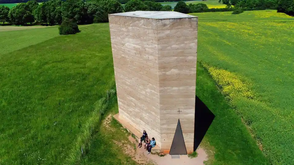 An aerial view shows three people sitting on the base of a tall, minimalist concrete structure in a lush green field with…