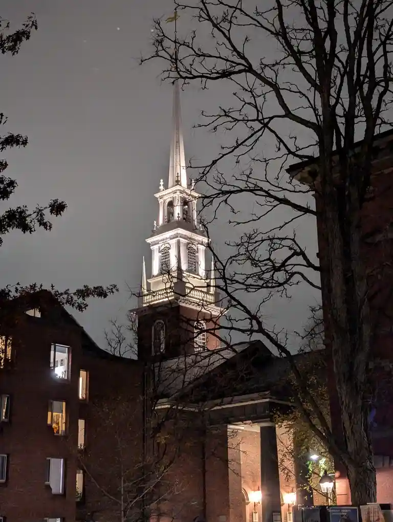 A nighttime photo of a historic church with a tall, illuminated steeple, framed by bare tree branches. The building is made…