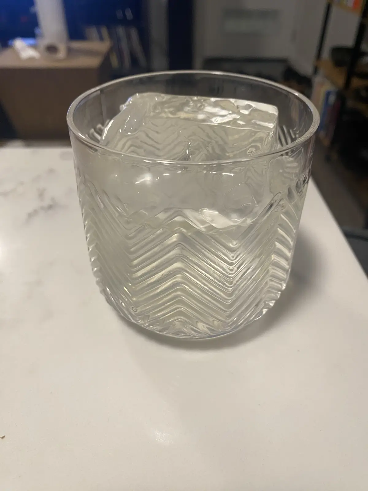 A clear glass with a zigzag pattern sits on a white countertop, containing a single large, clear ice cube. The overlaid text…