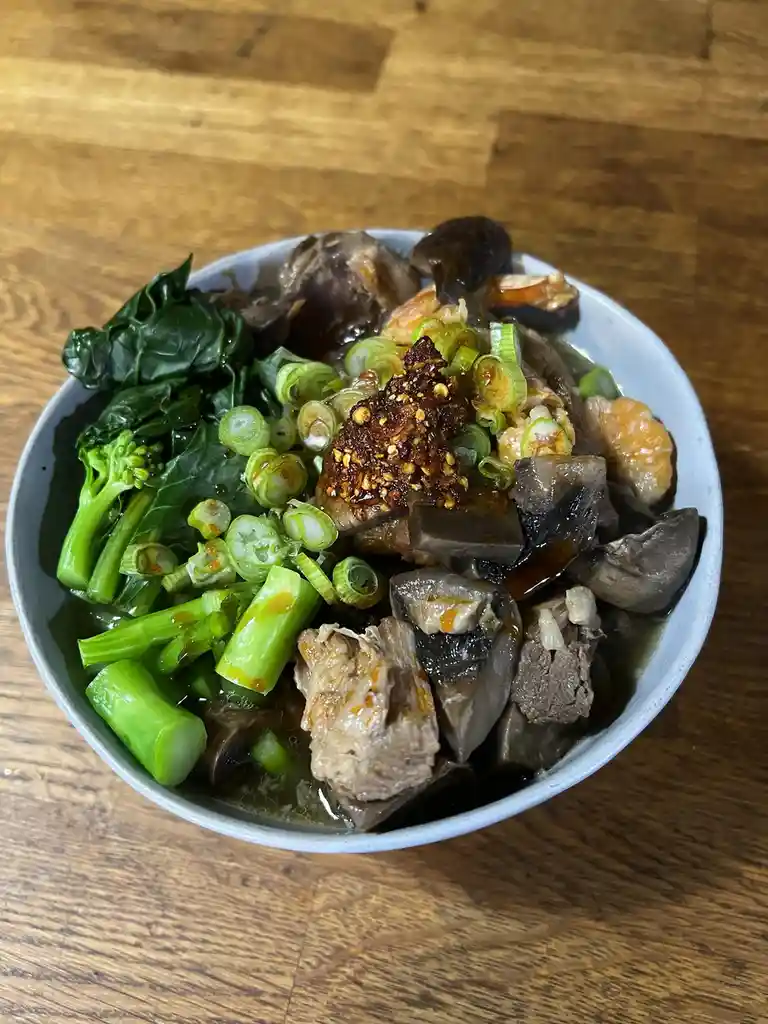 A close-up photo shows a light blue bowl filled with a dark broth, chunks of beef, dark mushrooms, green leafy vegetables…
