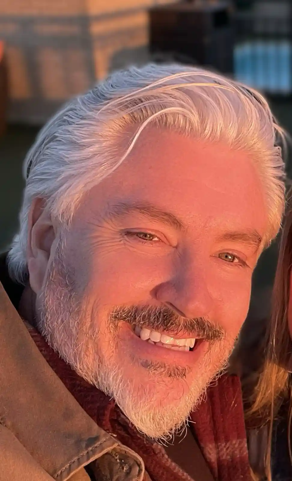 A close-up photo of a smiling man with white hair and a beard, wearing a brown jacket and red scarf, bathed in warm golden…
