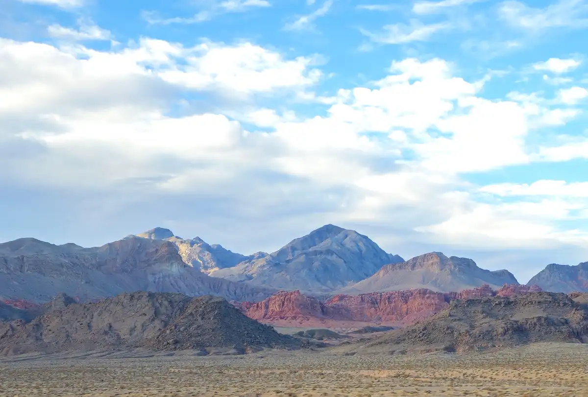 A scenic desert landscape featuring layered mountains with reddish and gray rock formations under a bright blue sky with…