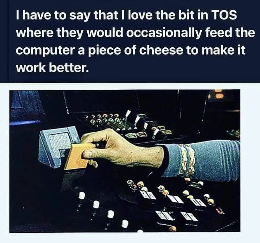 A hand in a blue sleeve holds a small piece of cheese and inserts it into a vintage computer terminal. The background shows…