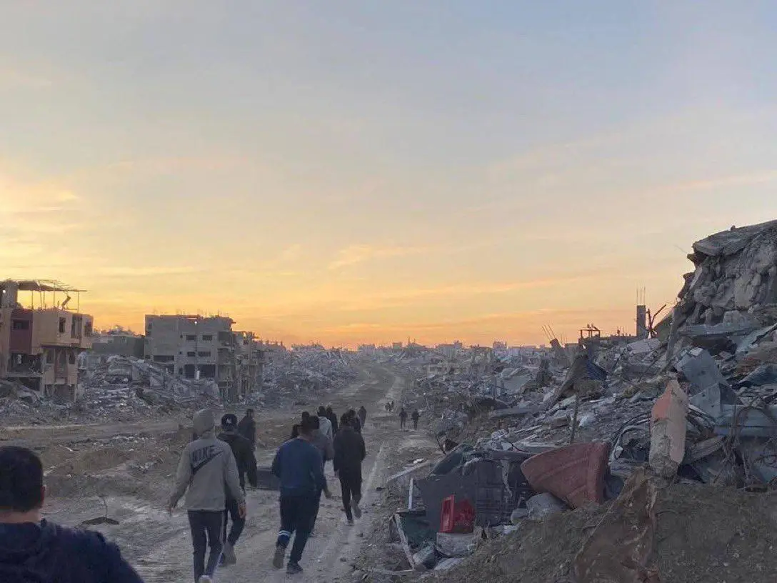 a group of people walk down a dirt path through a devastated urban area with rubble and destroyed buildings on both sides…